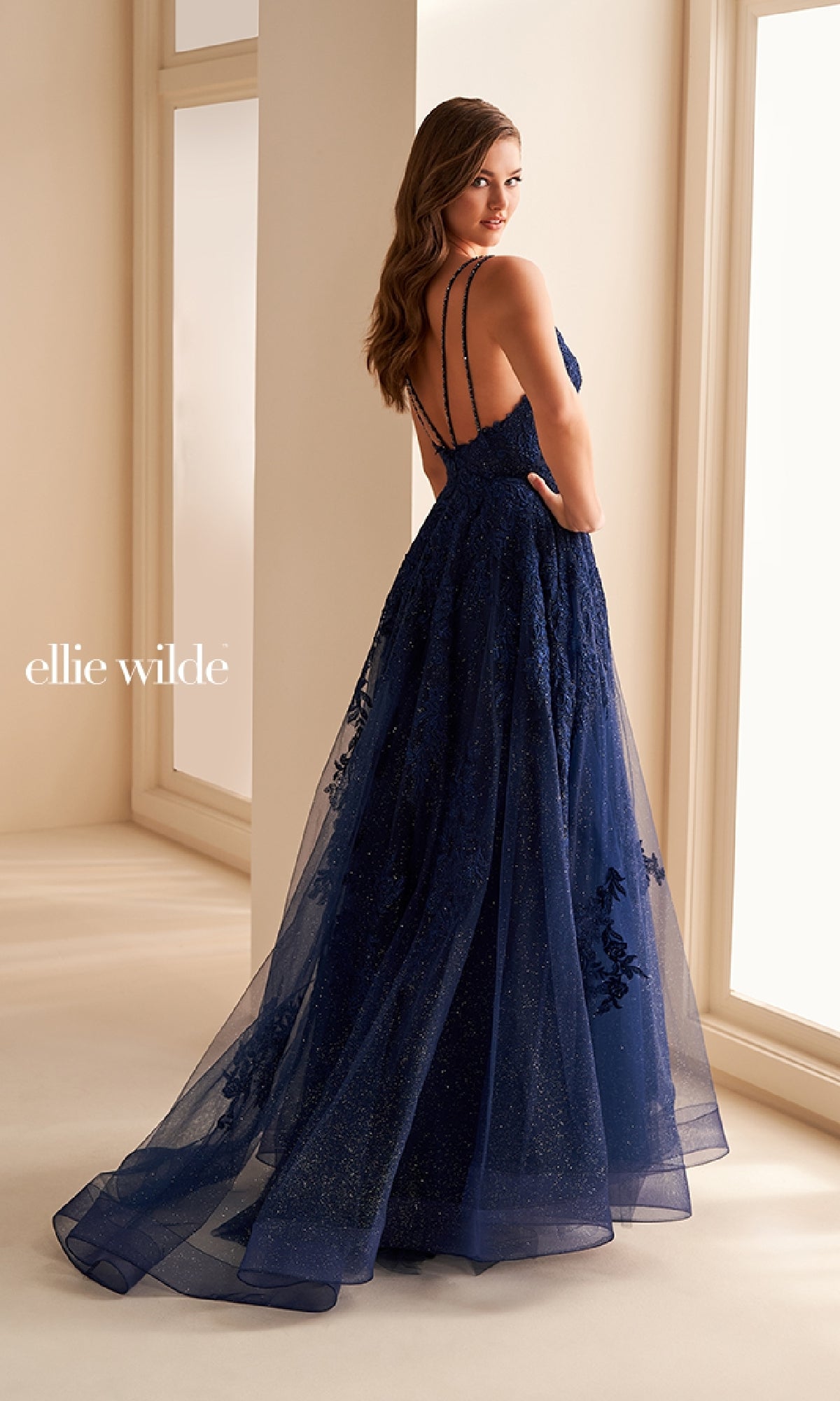 A woman in the Ellie Wilde EW36226 long prom dress—navy blue, A-line, with lace details, corset bodice, and sheer overlay—stands in a sunlit room facing away from the camera.